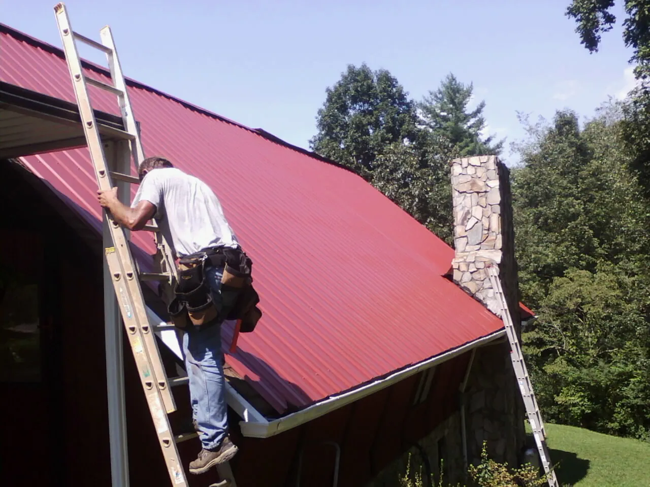 Roofer on a ladder beside a red metal roof and stone chimney — Soffit & Fascia Repair by Roofing Brothers in Dodge City